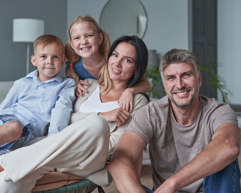 A family of four—two adults and two children—sit together on a couch, smiling at the camera in a living room.